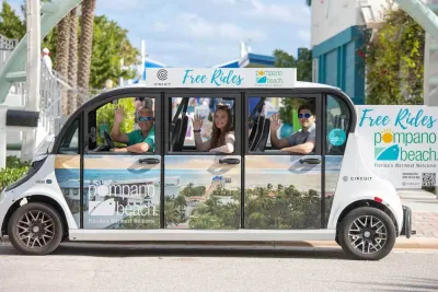 Small white microtransit shuttle with wrap depicting beach town in Pompano Beach, Florida with happy people waving from inside.