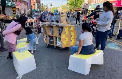 Children sit on temporary street furniture next to book cart at pop-up reading event at open streets event in Sunset Park in New York City.