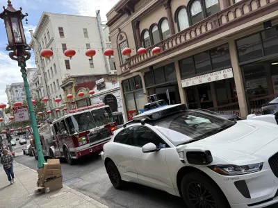 White Waymo car blocking street in front of red fire truck on narrow street with red Chinese lanterns in Chinatown, San Francisco, CA.