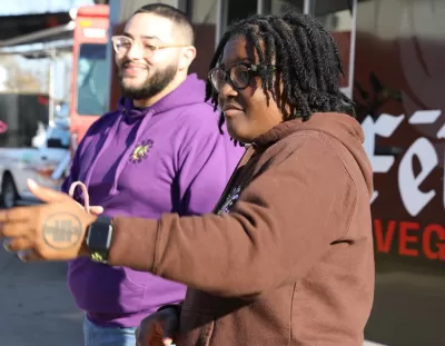Black woman in brown sweatershirt and Black man in purple sweatshirt stand in front of food truck.