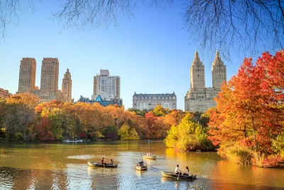View of lake in Central Park, New York City with people in rowboats and bright red and orange fall trees, buildings in background.
