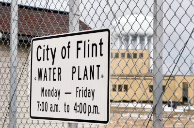 Close-up of sign on chain link fence "City of Flint Water Plant" with hours.