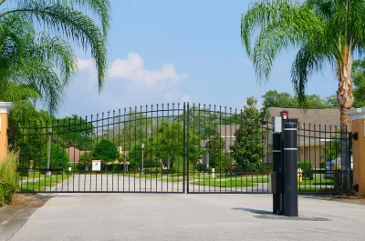 Closed black wrought iron gate in front of gated residential community with large palm trees along sides of street.