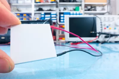 Close-up of hand holding small white square thermoelectric generator in lab with computer in background.