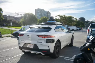 White Waymo autonomous vehicle in parking area in San Francisco, California with Ghirardelli vintage sign in background.