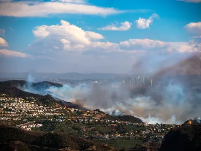 Heavy smoke from Palisades Fire burning in hillsides with homes with downtown Los Angeles visible in background.