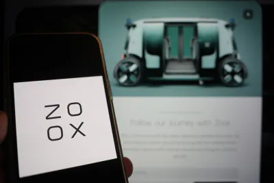 Close-up of smartphone with Zoox logo and screen with blurred image of Zoox autonomous vehicle in background.