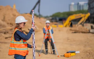 Woman and man in orange safety vests and hard hats doing surveying work at road construction site.