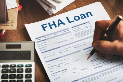Close-up of hand filing out FHA Loan application on desk with calculator next to paper.
