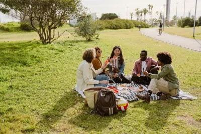 Group of five people sitting on blanket in park on sunny day having picnic.