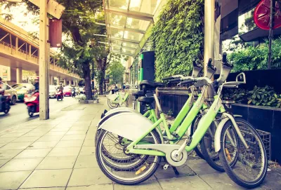 Green bike share bikes parked in a row on a commercial street with outdoor dining and greenery.