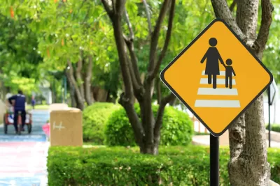 Yellow pedestrian crossing sign with black stick figures of mother and child crossing white crosswalk with blurred green trees on city street in background.