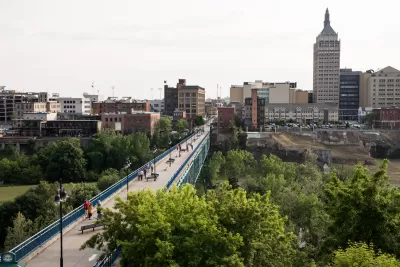 A long pedestrian bridge over a river in the High Falls District with skyline of Rochester, New York, in the backround.