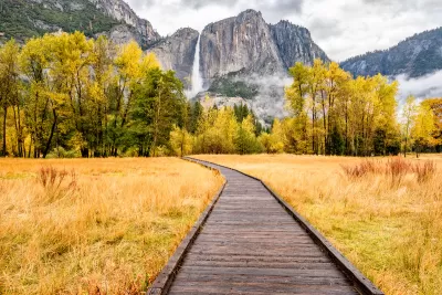 Wooden boardwalk through yellowed grass field with tall granite cliffs and waterfall in background in Yosemite National Park, California.