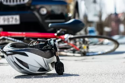 White bike helmet lies upturned on a road with red bicylce underneath the front of a far in the background.