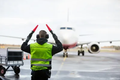 Ground crew person signaling to airplane with red batons on runway on a rainy day.