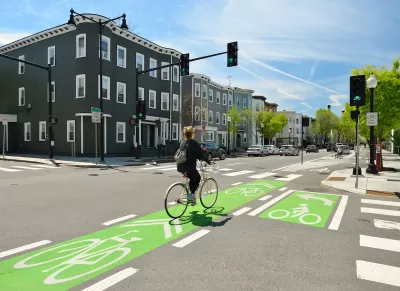 Cyclist on bike in green painted bike lane at intersection with three-story buildings in background.