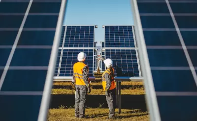 Two workers in white hard hats looking up at large photovoltaic solar panels.