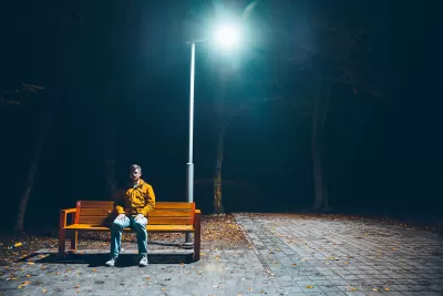 Man sitting on wooden bench under tall streetlight at night.