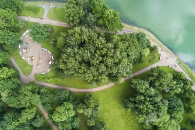 Bird's eye view of park with green trees, concrete walkways, and benches on lake or riverfront.