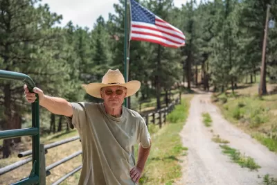 A cowboy rests his hand on a green gate on a sunny day in Colorado. The United States flag is behind him and he is wearing a straw cowboy hat and glasses. A dirt road is next to him.