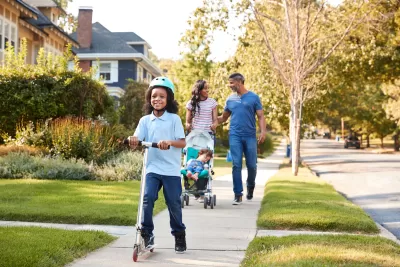 African-American parents push stroller down residential neighborhood sidewalk as young child rides scooter in front of them.