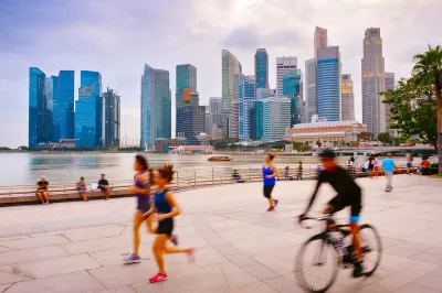 People running and cycling on wide paved path next to water with Singapore skyline with high-rises in background.