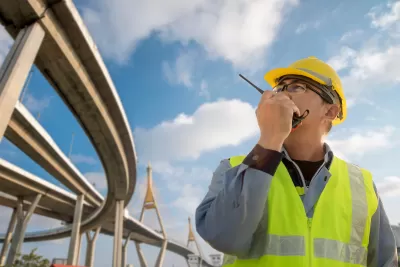 Civil engineer in yellow hard hat and safety vest talks on radio while looking up at freeway ramps.
