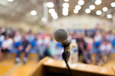 Close-up of microphone on speaker's dais with blurred crowd sitting in bright high school gym in background.