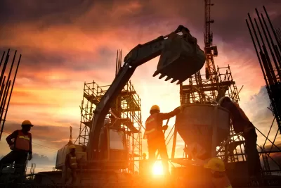 Construction workers, bulldozer, and unfinished bridge or highway sillhouetted against orange sunset sky