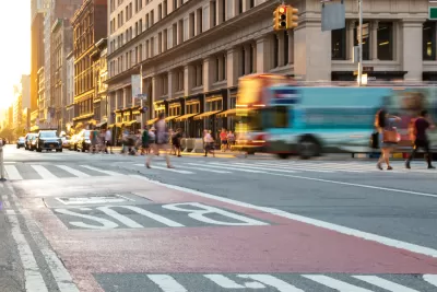 Low shot of red painted bus lane on New York City street with blurred bus, pedestrians, and buildings in background.