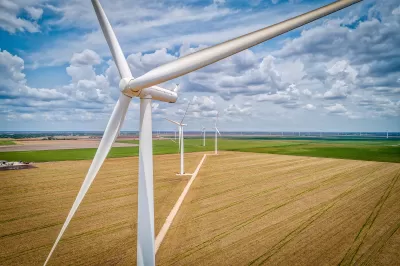 A line of white wind turbines surrounded by wheat and soybean fields with a cloudy blue sky in the background.