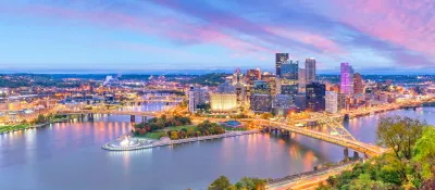 View of downtown Pittsburgh, PA with river and bridge in foreground at dusk.