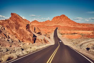 Two-lane highway with solid yellow center line and one car driving away against backdrop of red rock formations in Nevada.