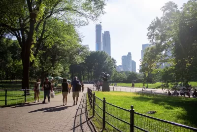 People walking on paved path in green city park with trees and tall city skyscrapers in background.