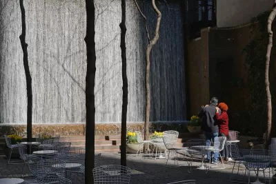 Man and woman watch waterfall in Paley Pocket Park in Manhattan, New York City. Park is filled with white tables and chairs.
