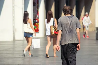Man walking down city sidewalk with sweat on back of his t-shirt on hot day.