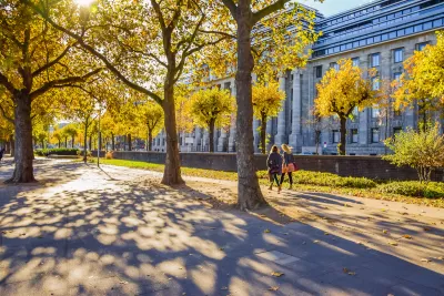 Two women walking on wide walkway among large trees next to neoclassical building in Cologne, Germany.