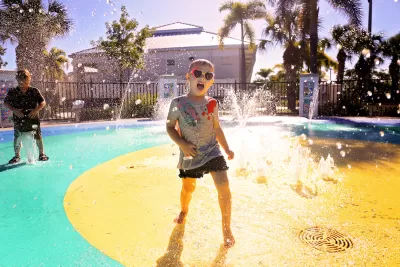 Child wearing heart-shaped sunglasses playing at splash pad in park.