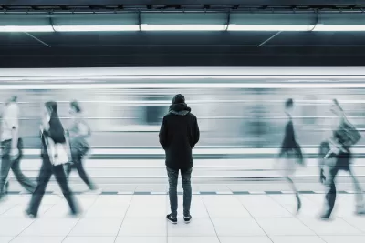 Monotone photo of man waiting on subway platform with blurry train passing by and other passengers walking by