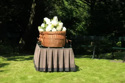 Woven willow casket with white flowers on top sits on table with pleated tablecloth on green lawn