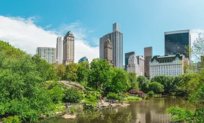 View of Central Park lake with people sitting on lakeside rocks and NYC high-rises in background.