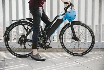 Close-up of black electric bike with person resting one foot on ground and holding blue helmet.