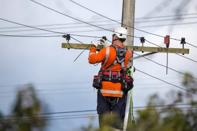 Worker in orange shirt and white helmet works at top of power line.