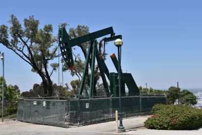 Green oil well pump jack surrounded by fence on a hill overlooking neighborhood.