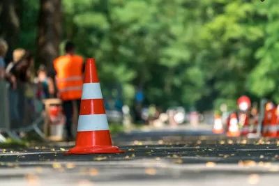 Close-up of orange and white traffic cone on street with blurred worker in orange vest in background.