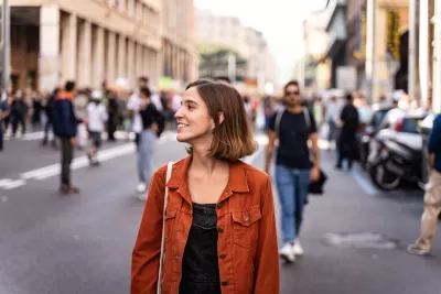 Brown-haired woman in an orange jacket walking down crowded street smiling