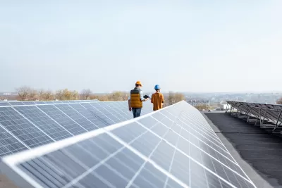 Rows of solar panels on a large rooftop with two workers in hard hats walking between rows.