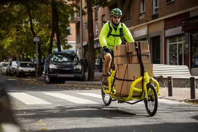 Man in bright yellow jacket riding yellow delivery cargo bike with several brown box packages tied down to bike on urban street.