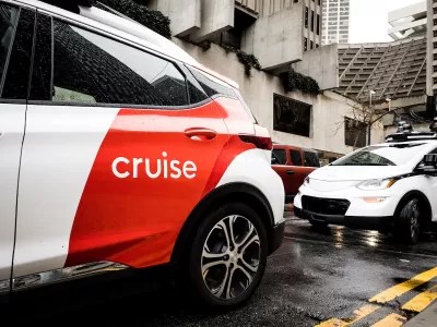 White and red Cruise autonomous car on San Francisco street with concrete Brutalist-style building in background on grey, rainy day.